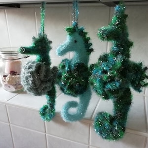 Pode incluir: Três ornamentos de cavalo-marinho de crochê pendurados em uma prateleira. Os cavalos-marinhos são feitos com fio verde, azul e prateado.