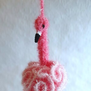 以下が含まれることがあります： ふわふわの質感の糸でできたピンクのフラミンゴのオーナメント。フラミンゴは長い首と脚、黒い目をしています。