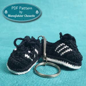 Peut inclure: Une paire de baskets en crochet noir et blanc avec des lacets blancs attachés à un porte-clés argenté. Les baskets ont trois bandes blanches de chaque côté.  PDF Pattern by Manufaktur Chouette.