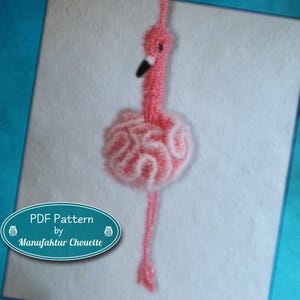 Könnte beinhalten: Ein gehäkelter Flamingo-Anhänger in Rosa mit strukturiertem, gerüschtem Körper und langem Hals. Der Flamingo hat einen schwarz-weißen Schnabel und ein kleines schwarzes Auge. Ein türkisfarbenes ovales Etikett zeigt den Text PDF Pattern by Manufaktur Chouette.