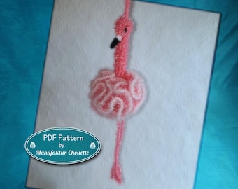 Op de afbeelding: Een roze gehaakt flamingo-ornament met een textuurrijk, geplooid lichaam en een lange nek. De flamingo heeft een zwart-witte snavel en een klein zwart oog. Een ovaal blauwgroen label in de hoek toont de tekst PDF Pattern by Manufaktur Chouette.