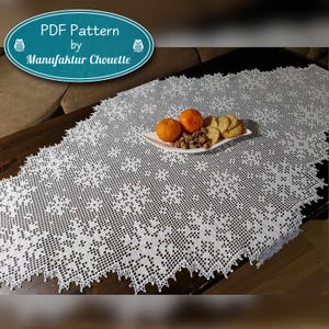 以下が含まれることがあります： 繊細な花柄の白いクロシェのテーブルランナー。ランナーは、オレンジとクッキーの入ったプレートが置かれた木製のテーブルの上に置かれています。