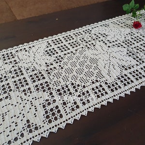 May include: White crocheted table runner with a delicate floral pattern. The runner is laid out on a dark brown wooden table.