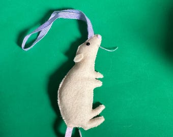 May include: A beige felt mouse with a purple felt tail and a purple felt loop for hanging. The mouse is on a green background.