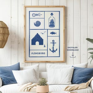 Puede incluir: Un estampado enmarcado con temática náutica en azul y blanco. El estampado presenta ilustraciones marítimas, como un barco en una botella y un ancla, con la palabra "FLENSBURG". El estampado cuelga sobre un sofá blanco con cojines azules.