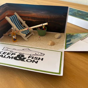 Könnte beinhalten: Handgefertigte Grußkarte mit einer Strandszene. Die Karte zeigt einen Miniatur-Strandstuhl, eine Angelrute und eine Bierflasche. Der Text "KEEP CALM & FISH ON" ist auf der Vorderseite aufgedruckt.