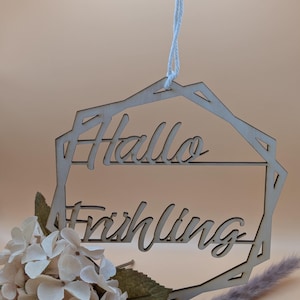 Peut inclure: Panneau en bois avec les mots "Hallo Frühling" découpés dans une police décorative. Le panneau est de forme géométrique et est suspendu par une ficelle blanche. Des fleurs blanches et de l'herbe séchée sont en bas.