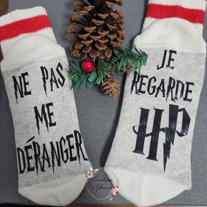 Peut inclure: Une paire de chaussettes grises avec des hauts blancs et des rayures rouges. L'une des chaussettes porte l'inscription "NE PAS ME DÉRANGER" en noir, l'autre "JE REGARDE HP". Une pomme de pin et de la verdure sont placées entre les chaussettes.
