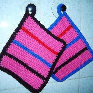 Könnte beinhalten: Zwei rosa gehäkelte Topflappen mit schwarzen, roten und blauen Streifen. Die Topflappen hängen an Saugnäpfen an einer gekachelten Wand.