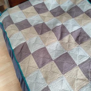 Könnte beinhalten: Eine handgefertigte Decke mit einem geometrischen Quadratmuster in Braun-, Beige- und Grautönen. Die Decke hat einen türkisfarbenen Rand. Die Strick- oder Häkelstruktur ist sichtbar, was auf einen gemütlichen, handgefertigten Artikel hindeutet.