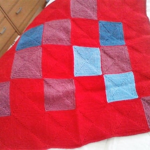 Könnte beinhalten: Eine handgestrickte rote Decke mit einem quadratischen Muster. Die Decke hat Quadrate in verschiedenen Farben, darunter Blau und Lila, die ein geometrisches Design bilden. Sie ist aus weichem Garn gefertigt und ideal zum Einkuscheln.