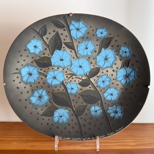 Könnte beinhalten: Ein dunkelgrauer Keramikteller mit einem blauen Blumendesign. Der Teller hat einen erhöhten Rand und eine strukturierte Oberfläche. Die Blumen sind stilisiert und haben einen zarten, fast skurrilen Look. Der Teller ist signiert mit "Helle Lucke Sundsvall" und "Ankie Frieland".