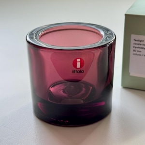 May include: A cylindrical dark purple glass tealight candle holder with a thick base and a red and white Iittala logo sticker on the front. Next to it is a light green box with text reading Tealight candle holder / Kynttilälyhty 60 mm.