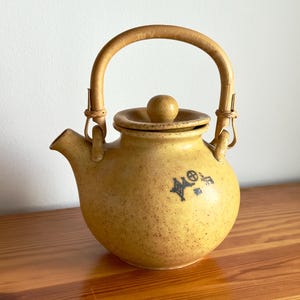 Peut inclure: Théière en céramique jaune-brun mouchetée, avec une anse et un couvercle en bambou. La théière a un bec verseur et un petit motif bleu foncé sur le côté. La poignée est arquée et fixée avec du matériau tissé. La théière est sur une surface en bois.