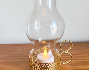 Puede incluir: Un portavelas de cristal transparente con una base y un asa de metal dorado. La base tiene un patrón decorativo de eslabones de cadena, y una pequeña vela que funciona con pilas en el interior. El portavelas está sobre una superficie de madera.
