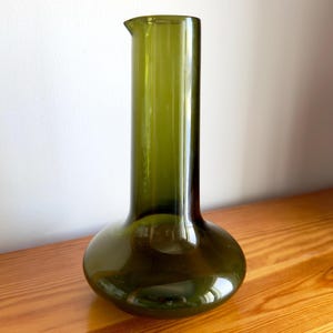 Peut inclure: Une carafe en verre vert olive, dotée d'une base large et arrondie qui se rétrécit en un col cylindrique élancé. Le récipient présente une finition lisse et translucide avec un petit bec verseur intégré au bord supérieur pour le service.