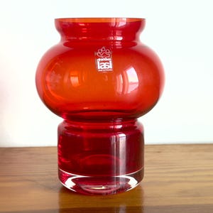 Könnte beinhalten: Eine leuchtend rote Glasvase mit einem abgerundeten, bauchigen Oberteil und einem zylindrischen Sockel. Die Vase hat einen durchsichtigen Aufkleber auf der Vorderseite mit einem Pfotenabdruck-Symbol und dem Text Riihimäen lasi MADE IN FINLAND.