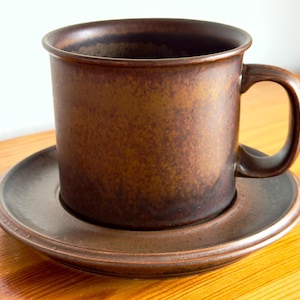 Peut inclure: Un ensemble tasse et soucoupe en céramique marron foncé présentant une finition texturée et mouchetée avec des nuances de marron plus sombre et de noir. La tasse possède un corps cylindrique avec une anse incurvée et un rebord plat.