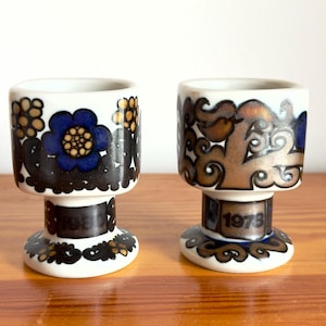 Op de afbeelding: Twee witte keramieken eierdopjes met donkerbruine en blauwe bloemmotieven. Het linker eierdopje toont het jaartal 1982, terwijl het rechter eierdopje het jaartal 1978 bevat. Beide items hebben een voetstuk en een cilindervormige kom.