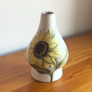 Op de afbeelding: Een witte keramische vaas met een taps toelopende hals en een ronde buik, voorzien van een handgeschilderd zonnebloemmotief in tinten geel, bruin en groen. Het bloemmotief loopt rond het onderste deel van de vaas en toont gedetailleerde bloemblaadjes en bladeren.
