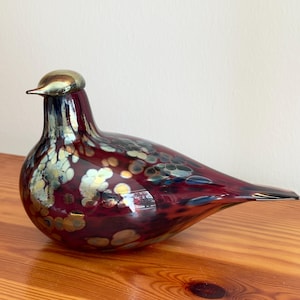 Może przedstawiać: Dekoracyjna szklana figurka ptaka w odcieniach bordowego, złotego i niebieskiego. Ptak ma zaokrąglony korpus i długi, spiczasty ogon. Powierzchnia jest nakrapiana złotymi i niebieskimi akcentami. Ptak siedzi na drewnianej powierzchni.