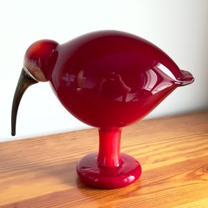 May include: A glossy, deep red glass bird sculpture with a long, curved beak. The bird has a rounded body and sits on a short, red pedestal base. The sculpture is displayed on a wooden surface.