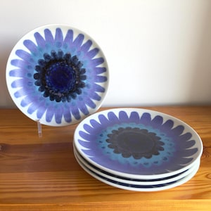 May include: A set of four round ceramic plates featuring a stylized floral pattern in shades of purple, blue, and black. The design consists of a dark central core surrounded by concentric rings of blue and purple petals radiating outward to the edge.