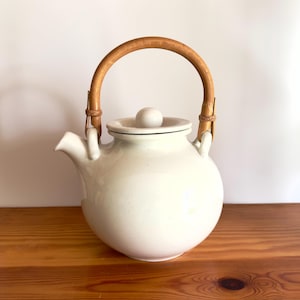 Op de afbeelding: Witte theepot van keramiek met een houten handvat en deksel. De theepot heeft een rond lichaam en een kleine tuit.