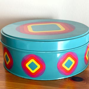 Peut inclure: Une boîte ronde turquoise avec un couvercle. Le couvercle et les côtés présentent un motif répétitif de carrés et de cercles concentriques colorés dans des tons de rouge, orange, jaune et violet. La boîte est posée sur une surface en bois.