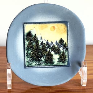 Puede incluir: Un plato redondo de cerámica gris con una escena de paisaje cuadrada y colorida. La escena representa un bosque de árboles verdes bajo un cielo amarillo con un sol. El plato se exhibe sobre un soporte transparente.