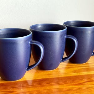 Könnte beinhalten: Drei marineblaue Keramikbecher mit gebogenen Griffen stehen auf einer Holzoberfläche. Die Becher haben eine matte Oberfläche und ein schlichtes, modernes Design. Die Becher sind leer und bereit, mit einem Getränk gefüllt zu werden.