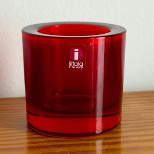 May include: A cylindrical, ruby-red glass candle holder with a thick base and a wide opening. The product has the "iittala FINLAND" logo on the front. The candle holder is sitting on a wooden surface.