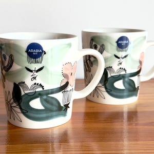 Peut inclure: Deux mugs en céramique blanche avec des motifs floraux verts, roses et noirs. Chaque mug a une anse et un autocollant circulaire bleu avec le texte "ARABIA 1873". Les mugs sont sur une surface en bois.