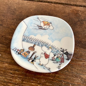Puede incluir: Un plato de cerámica blanco con una escena de invierno azul y blanco con un grupo de personajes Moomin. La escena incluye un personaje Moomin esquiando cuesta abajo por una colina nevada, otro personaje Moomin de pie en la colina y un tercer personaje Moomin de pie al lado de la colina. El plato está decorado con una escena de invierno azul y blanco.