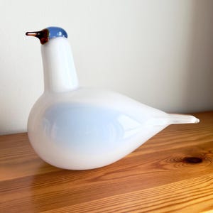 Könnte beinhalten: Eine weiße Glasvogel-Figur mit blauem Kopf und braunem Schnabel. Der Vogel hat einen runden Körper und einen langen Hals. Die Figur steht auf einer Holzoberfläche.