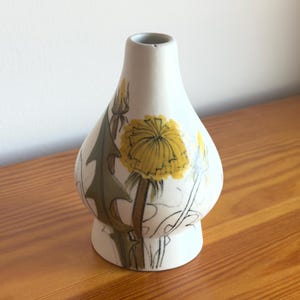 May include: A white ceramic vase with a tapered neck and a rounded body, featuring a hand-painted illustration of yellow dandelion flowers and green leaves. The design includes detailed line work and soft color shading on the exterior surface.