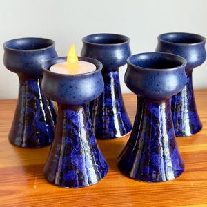 Könnte beinhalten: Fünf marineblaue Kerzenhalter aus Keramik mit floralem Design. Ein Halter hat eine brennende, batteriebetriebene Kerze. Die Halter haben einen ausgestellten Sockel und eine abgerundete Oberseite. Die Kerzenhalter stehen auf einer Holzoberfläche.
