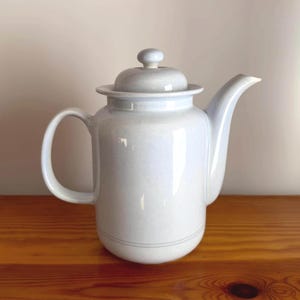 Puede incluir: Una cafetera de cerámica azul claro con tapa. La cafetera tiene un cuerpo redondeado y un pico en el lateral. La cafetera está sobre una superficie de madera.