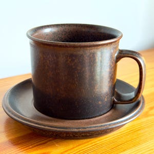 Könnte beinhalten: Ein dunkelbraunes Keramik-Tassenset mit passender Untertasse. Die Tasse hat einen zylindrischen Körper mit einem stabilen Henkel und eine gesprenkelte, melierte Glasuroberfläche. Die Untertasse hat einen leicht erhöhten Rand und die gleiche Textur.