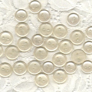 May include: A collection of 20 white plastic buttons with two holes each. The buttons are arranged in a random pattern on a white lace background.