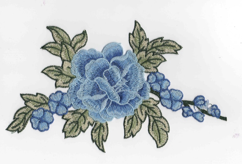 Op de afbeelding: Blauwe geborduurde bloemenapplicatie met groene bladeren. De bloem is een grote, enkele bloei met een gelaagd, gerimpeld ontwerp. De bladeren zijn gedetailleerd en realistisch.