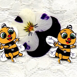 Puede incluir: Dos abejas de dibujos animados amarillas y negras con alas blancas y caras sonrientes. Las abejas están hechas de madera y tienen una apariencia de botón.