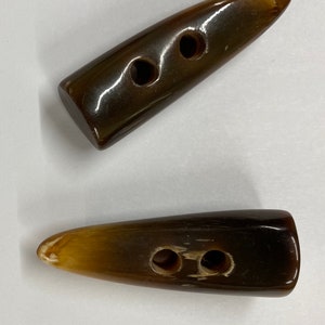 Könnte beinhalten: Zwei braune, hornförmige Knebelknöpfe mit je zwei Löchern. Die Knöpfe haben eine glänzende Oberfläche und eine Farbverlaufsfarbe, die von einem dunklen Braun zu einem helleren Hellbraun am spitzen Ende übergeht. Ideal für Mäntel oder Pullover.