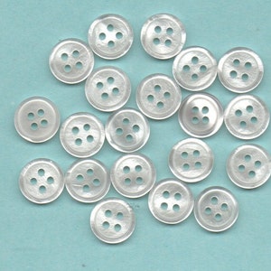 Può includere: Una collezione di 18 bottoni di plastica bianca, ognuno con quattro fori. I bottoni sono disposti in un cerchio su uno sfondo blu chiaro.