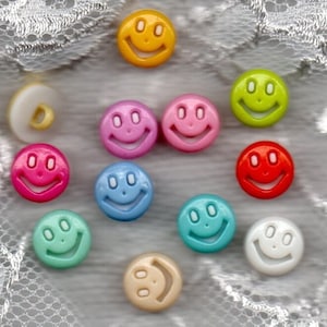Könnte beinhalten: Eine Sammlung bunter Smiley-Knöpfe in verschiedenen Farbtönen, darunter Gelb, Rosa, Grün, Rot, Blau und Weiß. Die Knöpfe haben eine glänzende Oberfläche und ein fröhliches Design. Ein weißer Knopf mit einer Schlaufe ist ebenfalls vorhanden.