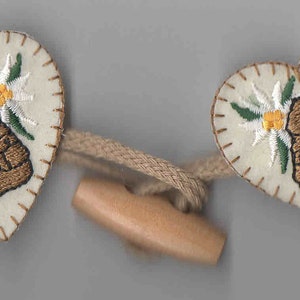 May include: Two white felt heart-shaped buttons with brown embroidered backpacks and white flowers. The buttons are connected by a brown cord with a wooden toggle.
