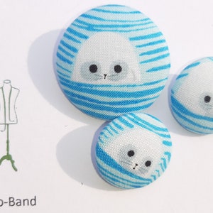 May include: Three fabric buttons with a blue and white striped pattern and a white seal face design. The buttons are different sizes.