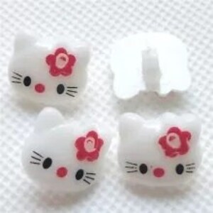 Könnte beinhalten: Vier weiße Plastikknöpfe mit einem Cartoon-Katzenkopf-Design. Jeder Knopf hat eine rosa Blume und schwarze Augen.