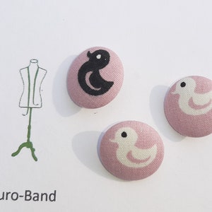May include: Three fabric buttons with a pink background and a white or black duck design. The text "Euro-Band" is printed below the buttons.