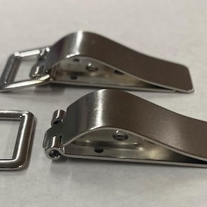 Puede incluir: Dos hebillas metálicas plateadas con un anillo rectangular y un diseño de palanca curvada. Las hebillas probablemente sirven para asegurar correas o cierres en bolsos, cinturones u otros accesorios.
